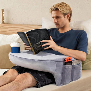 Coussin de lecture avec rangement latéral pratique posé sur les jambes d’une personne assise sur un lit, avec compartiments contenant une télécommande et un téléphone, et une boisson posée dessus.