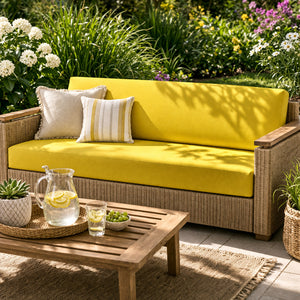 Un coussin salon de jardin imperméable rectangulaire posé sur un canapé extérieur en résine tressée, accompagné de coussins décoratifs, dans un jardin verdoyant avec une table basse en bois et une carafe citronnée.