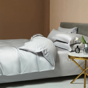 Une parure de lit luxe en satin gris clair avec finition lisse et brillante, disposée dans une chambre moderne avec table de chevet dorée et ambiance élégante.