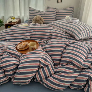Parure de lit rayure gaze de coton double couche aux tons bleus et rosés, avec couette moelleuse et coussins assortis, posée dans une chambre lumineuse avec un plateau petit-déjeuner sur le lit.