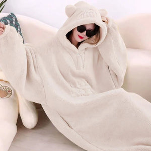Une personne assise sur un canapé porte un plaid avec manches à capuche au tissu duveteux, avec de larges manches et une capuche à petites oreilles, créant une tenue cocooning idéale pour se détendre à la maison.