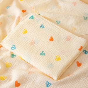Un plaid bébé en gaze de coton gaufrée, délicatement plié, avec de petits motifs en forme de cœurs, posé sur un tissu assorti créant une ambiance douce et apaisante.