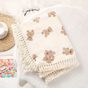 Un plaid bébé à motif oursons avec franges délicates, posé sur un fauteuil moelleux, accompagné d’un livre et d’objets doux dans une ambiance chaleureuse et apaisante.