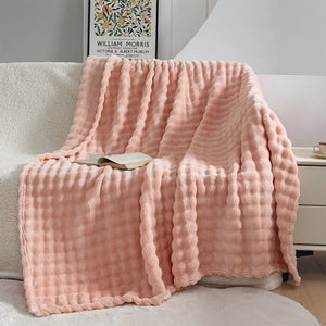 Un plaid fausse fourrure doux et moelleux à texture gaufrée, délicatement drapé sur un fauteuil avec un livre posé dessus, créant une atmosphère chaleureuse et invitante dans un salon lumineux.