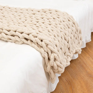 Un plaid grosse maille beige épais et moelleux, délicatement posé sur le bord d’un lit blanc, créant une ambiance chaleureuse et cocooning dans une chambre lumineuse.