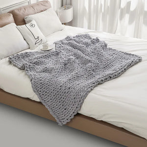 Un plaid grosse maille style scandinave drapé sur un lit, avec de larges tresses épaisses et un rendu moelleux, créant une atmosphère chaleureuse et cosy dans une chambre lumineuse.