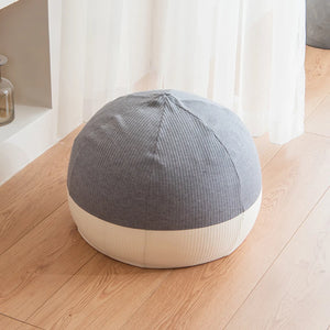 Un pouf de salon respirant en tissu texturé gris et beige, de forme arrondie, posé sur un sol en bois clair près de rideaux blancs, dans une ambiance minimaliste et lumineuse.
