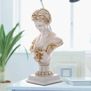 Buste de Vénus en finition dorée antique sur bureau blanc près d'une lampe et plantes