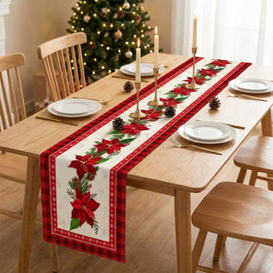 Chemin de table de Noël à motif poinsettias rouges et bordure à carreaux posé sur une table en bois dressée avec assiettes blanches, bougies dorées et sapin illuminé en fond