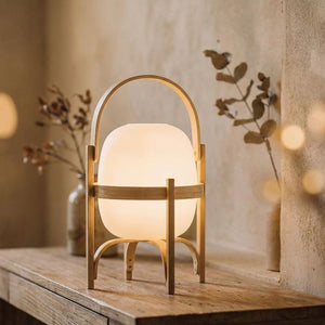 Lampe lanterne en bois clair avec globe de verre opale allumée posée sur une console dans une pièce chaleureuse