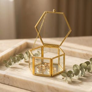 Boîte à bijoux hexagonale en verre transparent et laiton doré posée sur un plateau en marbre avec des bagues