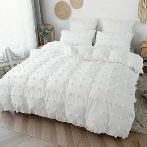 Parure de lit blanche à pompons vue de profil dans une chambre lumineuse avec chaise en rotin