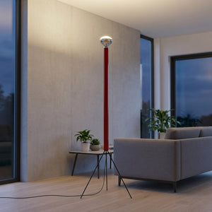 Lampadaire trépied rouge allumé près d'un canapé devant de grandes fenêtres au crépuscule