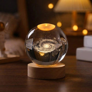 Boule de cristal gravée en 3D représentant une galaxie éclairée d'une lumière chaude sur socle en bois