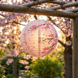 Lanterne sphérique rose aux motifs sakura suspendue sous une pergola bambou dans un jardin fleuri