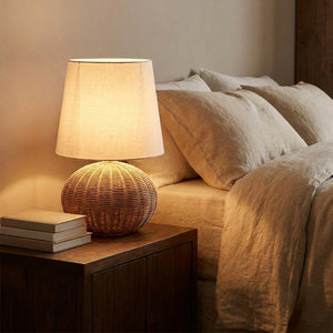 Lampe en rotin tressé allumée sur une table de chevet en bois près d un lit en lin beige