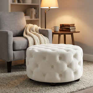 Tabouret pouf blanc en velours utilisé comme repose-pieds près d'un fauteuil gris