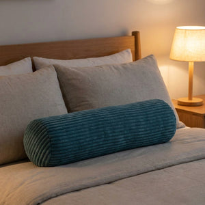 Traversin en velours côtelé bleu foncé posé sur un lit devant les oreillers avec une lampe de chevet allumée