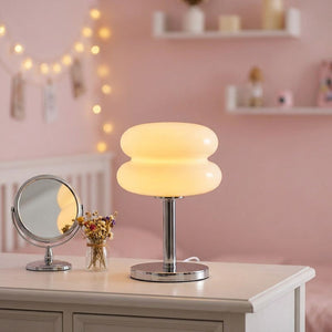 Lampe de chevet en verre forme macaron allumée sur un bureau dans une chambre pastel avec un miroir