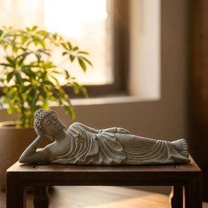 Statue de Bouddha couché en grès sur une table en bois près d'une plante verte en lumière dorée