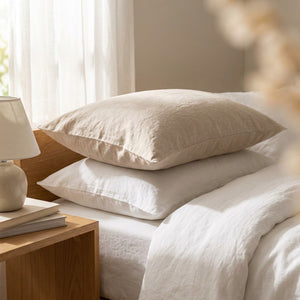 Taies d'oreiller en lin beige et blanc empilées sur un lit près d'une fenêtre