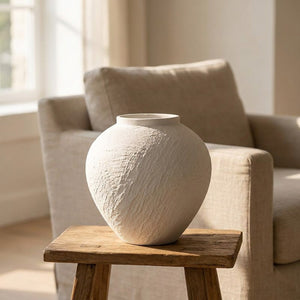 Vase en céramique blanche posé sur un guéridon devant un fauteuil en lin