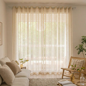 Voilage tulle beige habillant une baie vitrée dans un salon cosy avec canapé et fauteuil en rotin
