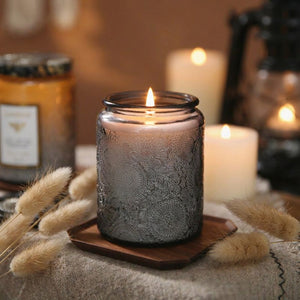 Bougie parfumée en pot en verre gris allumée sur plateau bois avec herbes séchées et éclairage ambiant