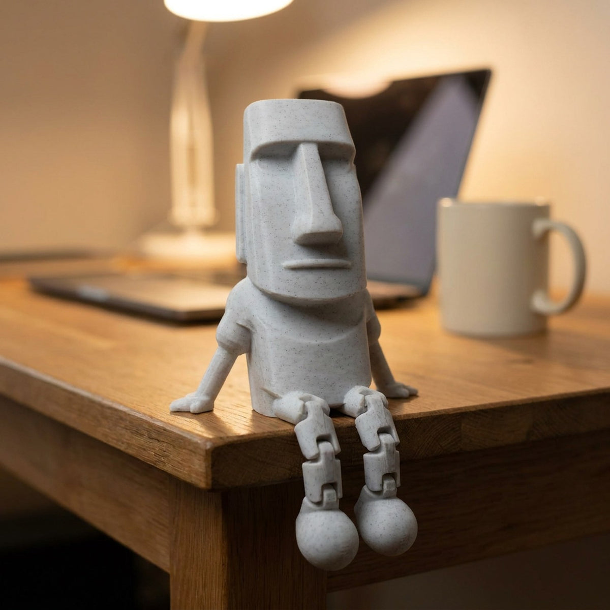 Figurine Moai grise posée au bord d'un bureau les jambes articulées pendantes près d'un ordinateur