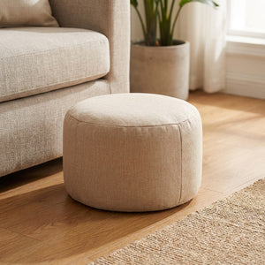 Pouf tabouret rond en lin beige posé au sol dans un salon lumineux près d'un fauteuil