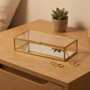 Boîte à bijoux en verre transparent et cadre doré posée sur une table de nuit en bois avec petite plante