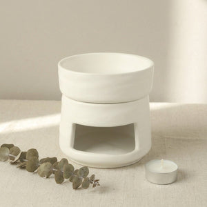 Brûle-parfums en céramique mate blanche posé sur surface en lin avec brindilles d'eucalyptus