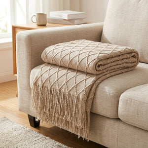 Plaid tricoté beige plié sur un canapé en lin montrant sa maille losange et ses franges