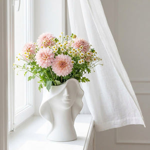 Vase sculpté blanc avec dahlias roses et marguerites sur rebord de fenêtre, lumière matinale douce