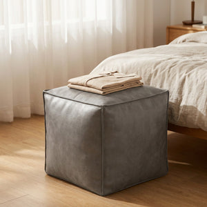 Pouf cube en cuir gris avec vêtements pliés posés dessus dans une chambre lumineuse