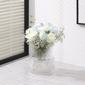 Vase en verre rayé avec roses bleues et blanches près d'une fenêtre à store blanc