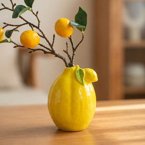Vase en céramique jaune en forme de citron garni de branches fleuries sur une table en bois