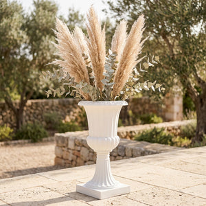 Vase médicis blanc posé sur une terrasse en pierre avec des herbes de pampa dans un jardin méditerranéen
