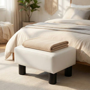 Tabouret repose-pieds blanc au pied d'un lit avec un plaid beige plié posé dessus dans une chambre lumineuse