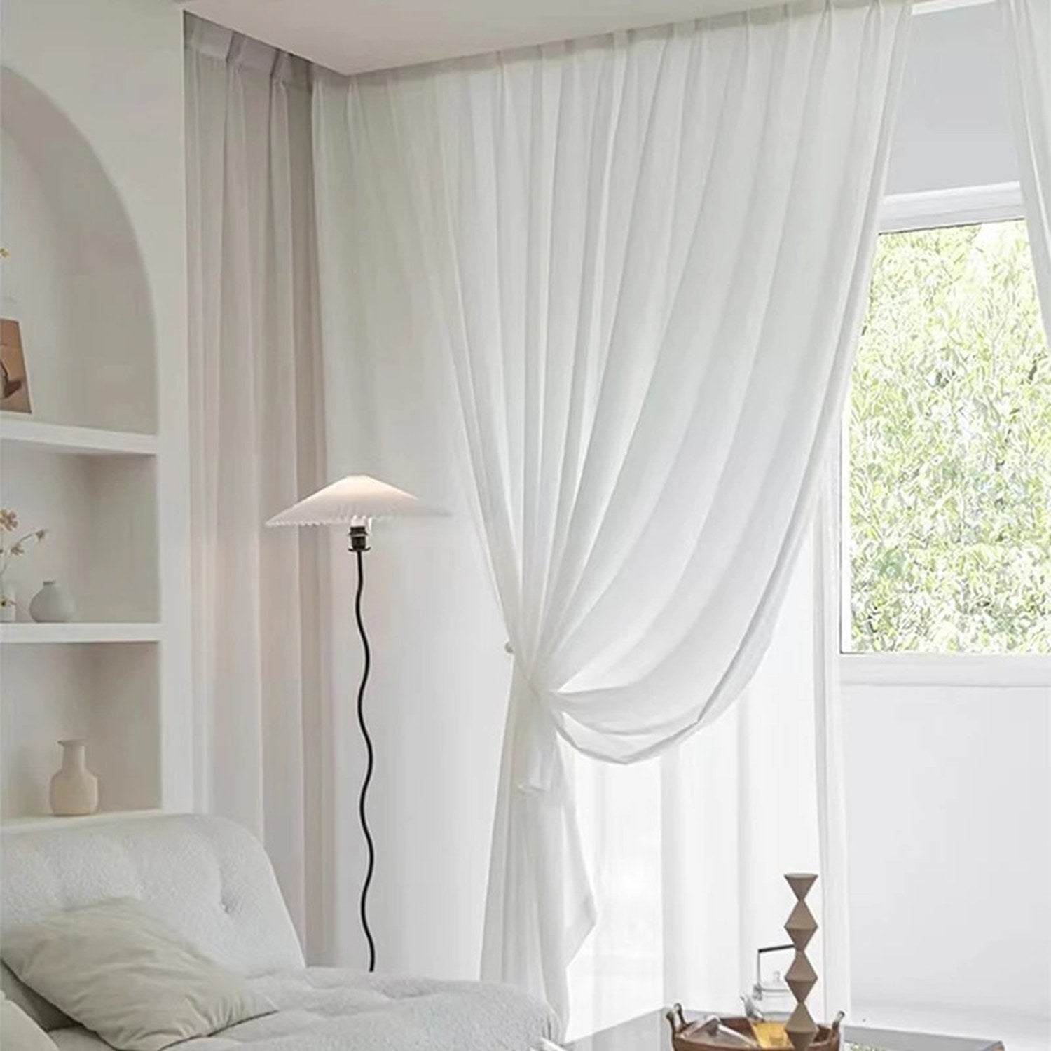 Voilage blanc suspendu à une tringle plafond dans un salon lumineux avec lampadaire et niche murale arrondie