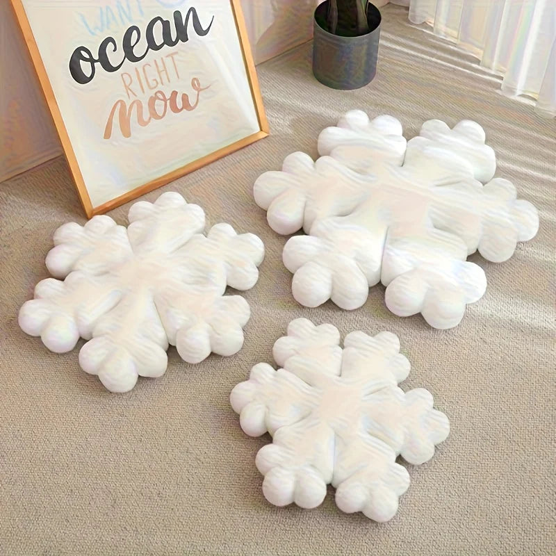 Des coussins Noël doux en forme de flocon de neige posés sur un tapis, avec un cadre décoratif et une plante en arrière-plan, créant une ambiance chaleureuse et apaisante.