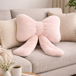 Coussin canapé moelleux en forme de nœud, de couleur rose poudré, posé au centre d’un canapé beige avec d’autres coussins clairs dans un salon lumineux et cosy.