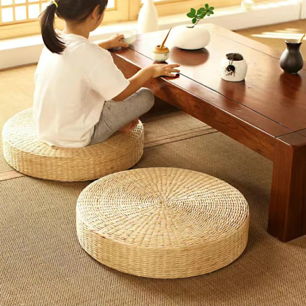 Coussin de sol en paille style japonais, rond et tressé, placé sur un tapis beige devant une table basse en bois, créant une ambiance zen et naturelle.