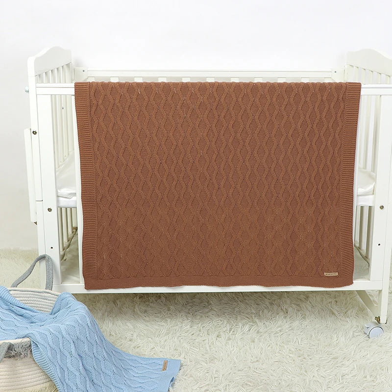 Une couverture bébé cocooning en coton naturel avec motif texturé, soigneusement posée sur un berceau blanc dans une chambre douce et lumineuse.