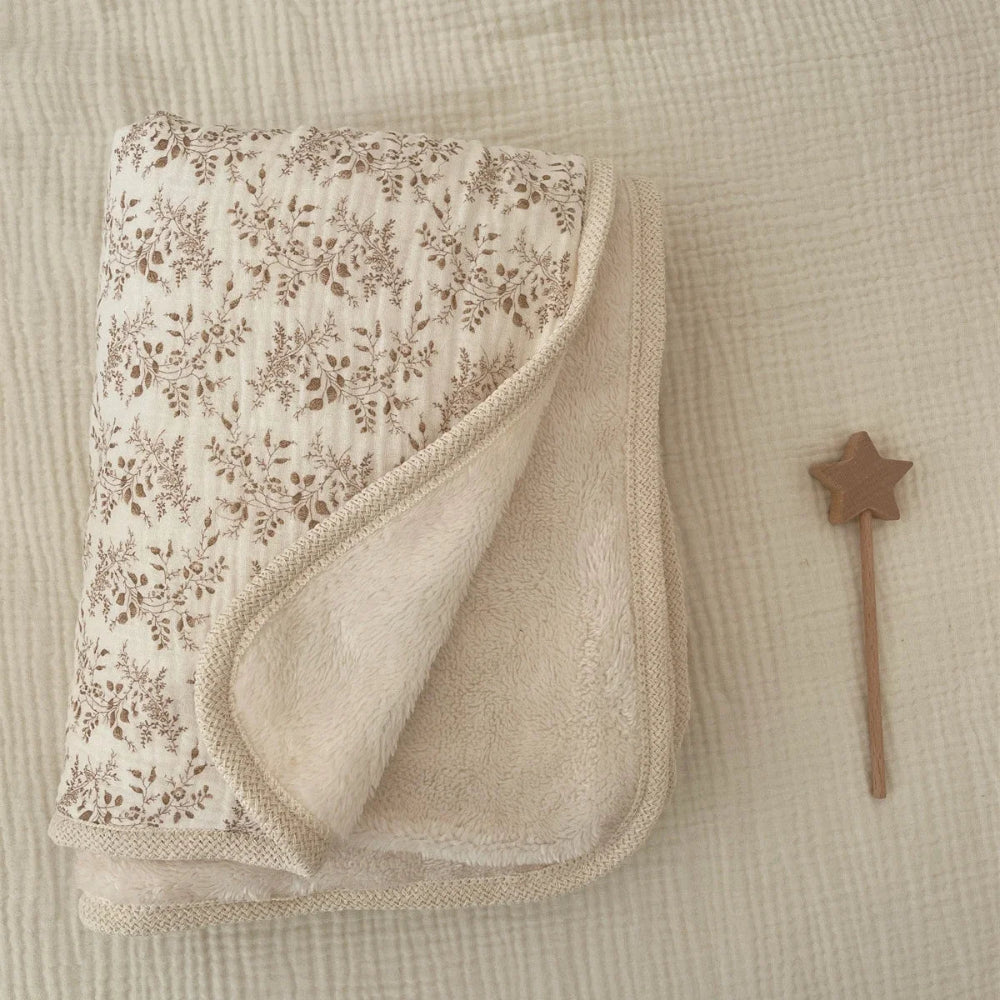 Une couverture bébé réversible avec une face à motif floral délicat et une face en tissu doux, délicatement pliée sur un textile gaufré avec un petit accessoire en bois à côté.