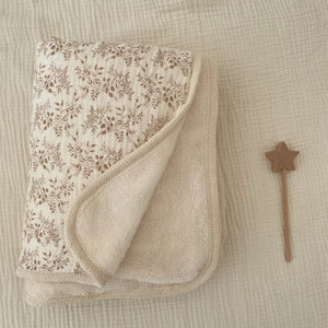 Une couverture bébé réversible avec une face à motif floral délicat et une face en tissu doux, délicatement pliée sur un textile gaufré avec un petit accessoire en bois à côté.
