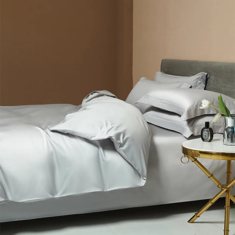 Une parure de lit luxe en satin gris clair avec finition lisse et brillante, disposée dans une chambre moderne avec table de chevet dorée et ambiance élégante.