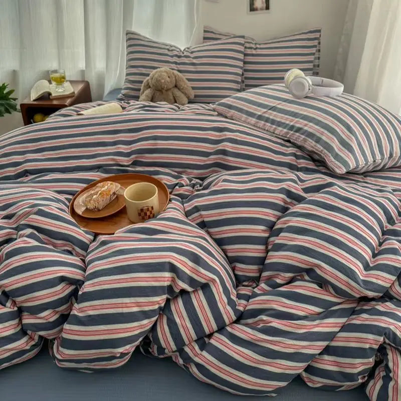 Parure de lit rayure gaze de coton double couche aux tons bleus et rosés, avec couette moelleuse et coussins assortis, posée dans une chambre lumineuse avec un plateau petit-déjeuner sur le lit.