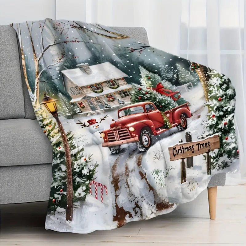 Un plaid chaud au motif vintage esprit Noël illustrant une scène enneigée avec une maison décorée et un camion ancien transportant un sapin, posé sur un canapé dans un salon lumineux.