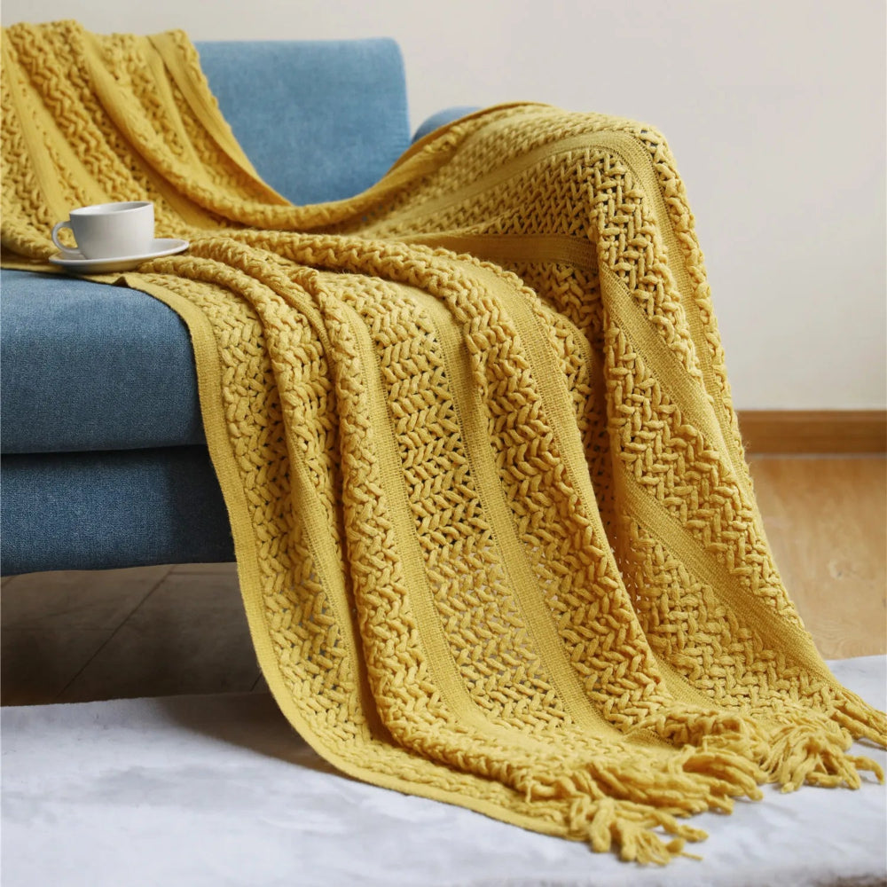 Un plaid crochet bohème beige avec franges élégantes, délicatement posé sur un fauteuil en rotin clair, accompagné d’un coussin neutre dans une ambiance douce et naturelle.